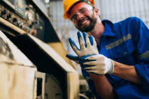 A man in a blue work uniform, yellow hard hat, and safety glasses, wearing protective gloves with blue palms, is smiling while working in an industrial setting. He stands next to machinery, appearing to be adjusting his gloves or preparing for a task.