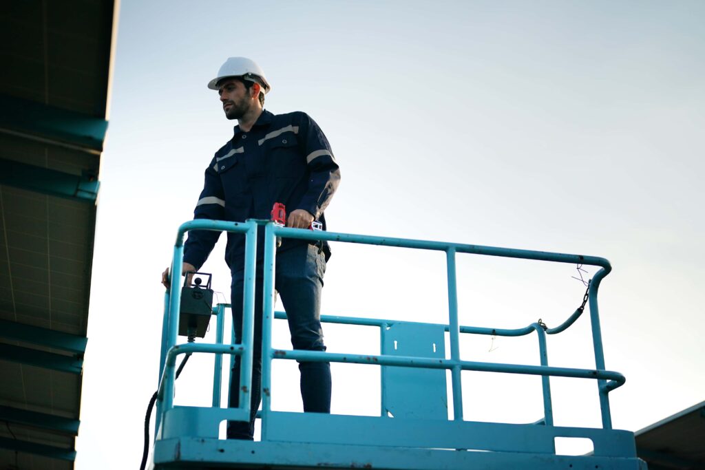 A man wearing a white hard hat and dark work clothes stands on an elevated platform, operating machinery or controls. The background shows a clear sky, and the man is focused on his task, ensuring safety and compliance.