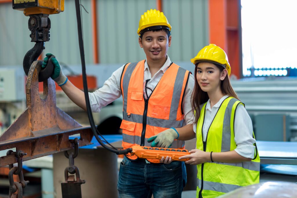 Two construction workers in high-visibility vests and yellow helmets stand in an industrial setting. One operates an auto draft control panel while the other steadies a hoisting mechanism. Both appear focused and alert, demonstrating teamwork and safety in their environment.