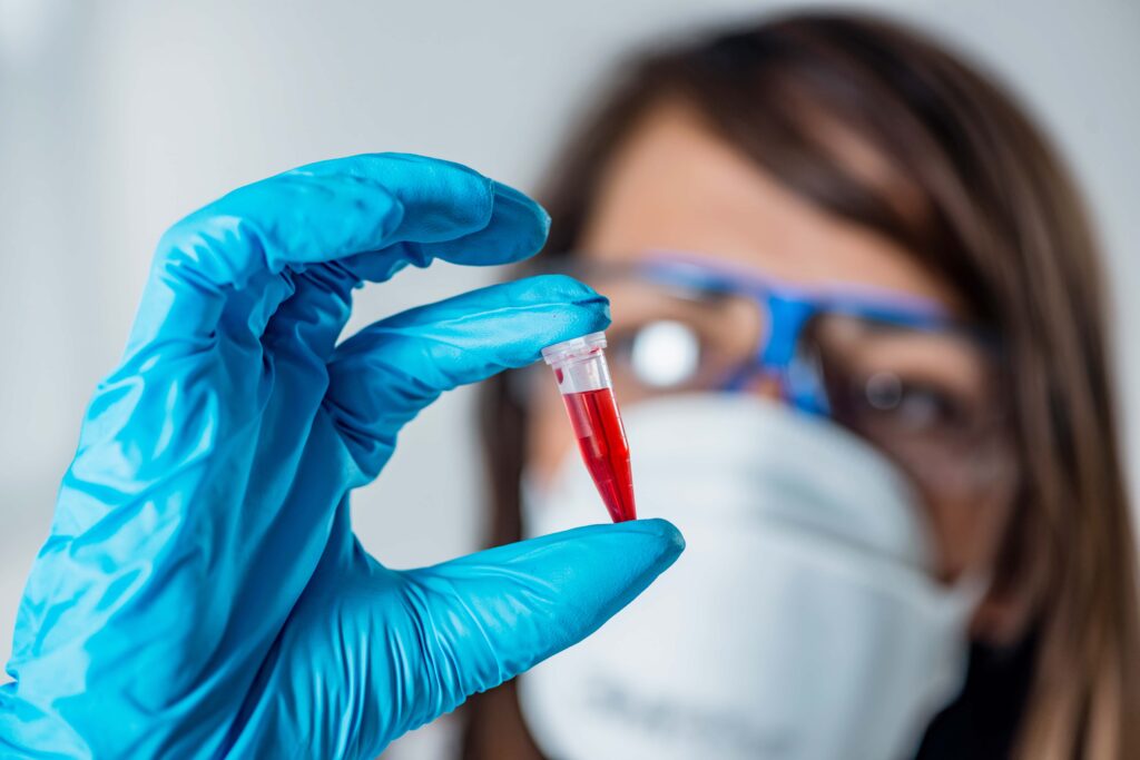 A person wearing blue gloves, safety glasses, and a face mask holds up a small vial containing a red liquid, emphasizing the importance of workplace safety. The focus is on the vial, essential for bloodborne pathogens training, while the person’s face is slightly blurred in the background.