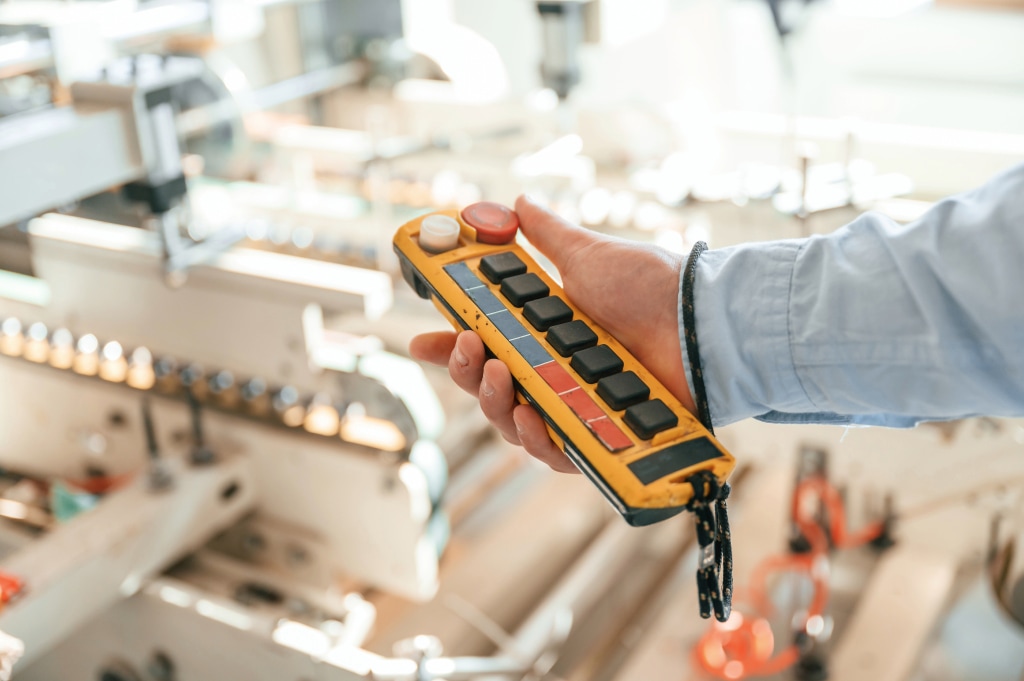 A person holds a yellow industrial remote control with multiple black buttons and a red emergency stop button, ensuring worker safety. The background is blurred, showing a machine with metallic components designed for productivity technology.