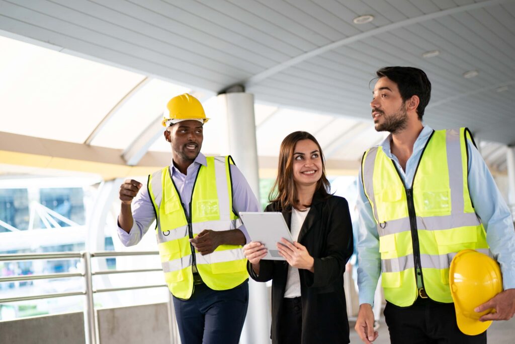 Three people walking together in a modern building. Two of them are in construction attire, wearing yellow safety vests and helmets. The third person, a woman in business attire, is holding a tablet. They appear to be engaged in a discussion about workplace safety insights.