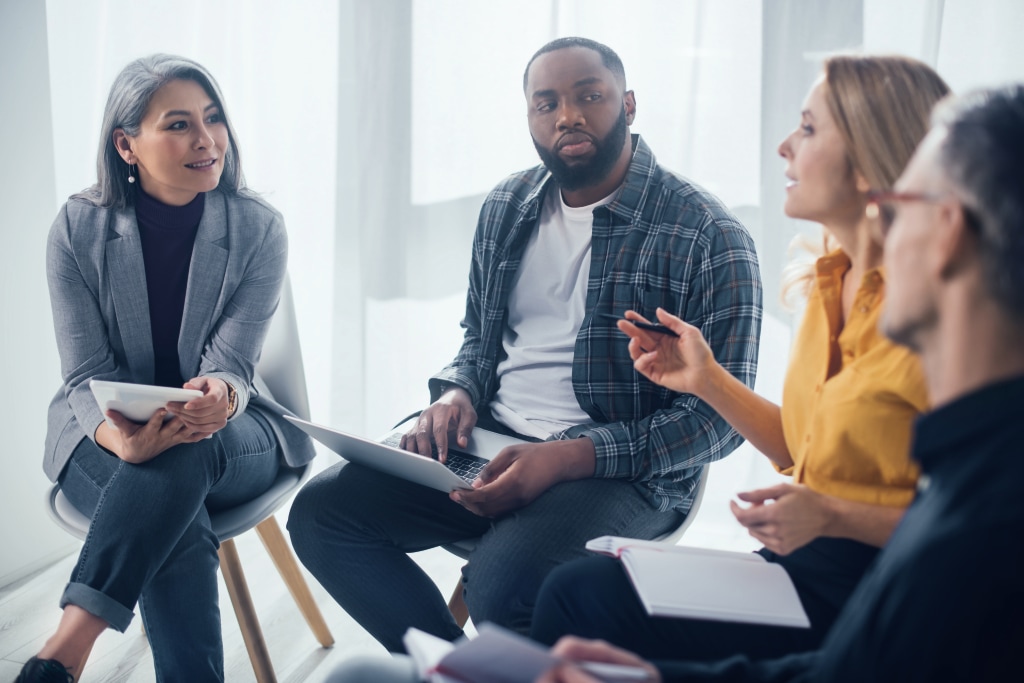 A diverse group of four people sits in a circle, deeply engaged in discussion about employee mental health. One person gestures while speaking, as others listen attentively, with notebooks and a laptop in hand. The setting is bright with natural light, offering warmth even during the cold season.