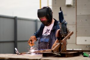 A person, ensuring cutting safety, wears a welding mask and gloves while working outdoors with a welding tool, producing bright sparks. They stand at a wooden table scattered with metal pieces and equipment. In the background, there's a metal fence and nearby buildings.