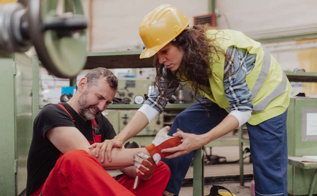 A woman in a yellow safety vest and hard hat tends to a man in red overalls who is holding his injured arm in a workshop. Demonstrating effective first aid, she addresses the workplace hand injury while various tools and machinery can be seen in the background. The man appears to be in pain.