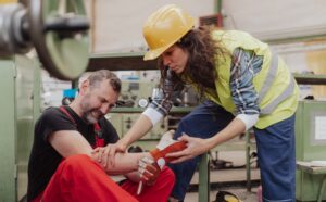 A woman in a yellow safety vest and hard hat tends to a man in red overalls who is holding his injured arm in a workshop. Demonstrating effective first aid, she addresses the workplace hand injury while various tools and machinery can be seen in the background. The man appears to be in pain.