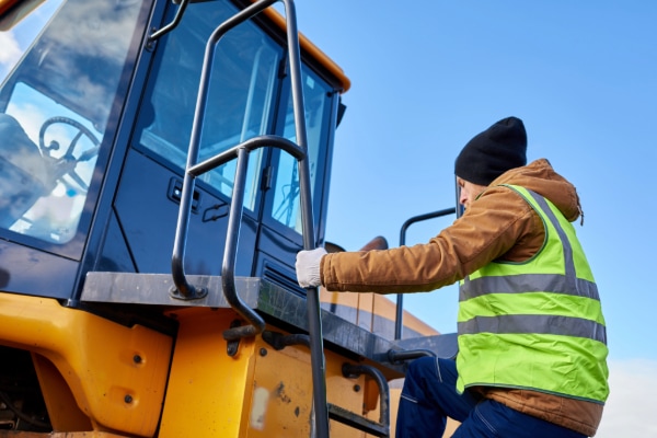 A person wearing a high-visibility vest and beanie climbs onto a yellow construction vehicle under a clear blue sky, embracing winter training in industrial workplaces. The individual holds onto the metallic handrail while ascending the steps to the cabin.