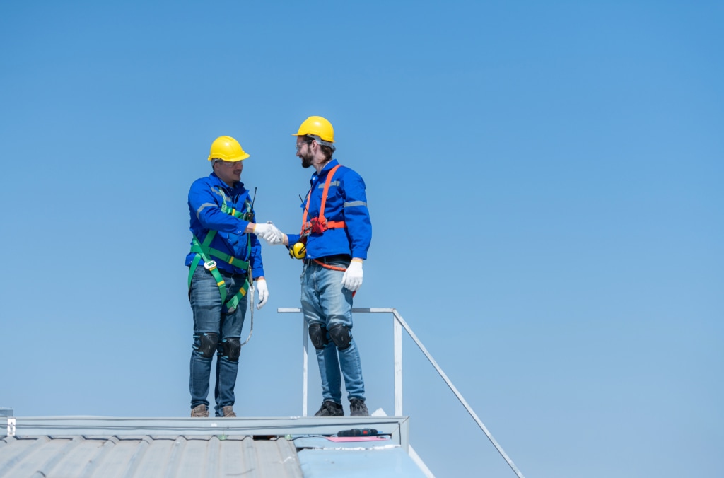 Two construction workers in blue jackets and yellow hard hats shake hands on a rooftop, both secured with safety harnesses as part of their fall protection gear. The sky is clear and blue in the background.