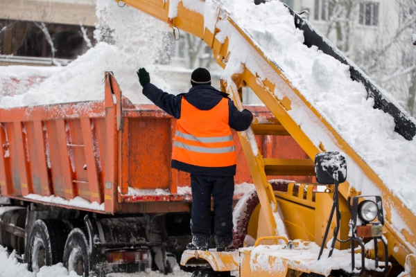 A worker in an orange reflective vest efficiently manages snow removal, using a large machine to load snow into a dump truck. Despite the falling snow and chilly temperatures, their focus on winter wellness ensures they stay warm and safe in the snowy urban environment.