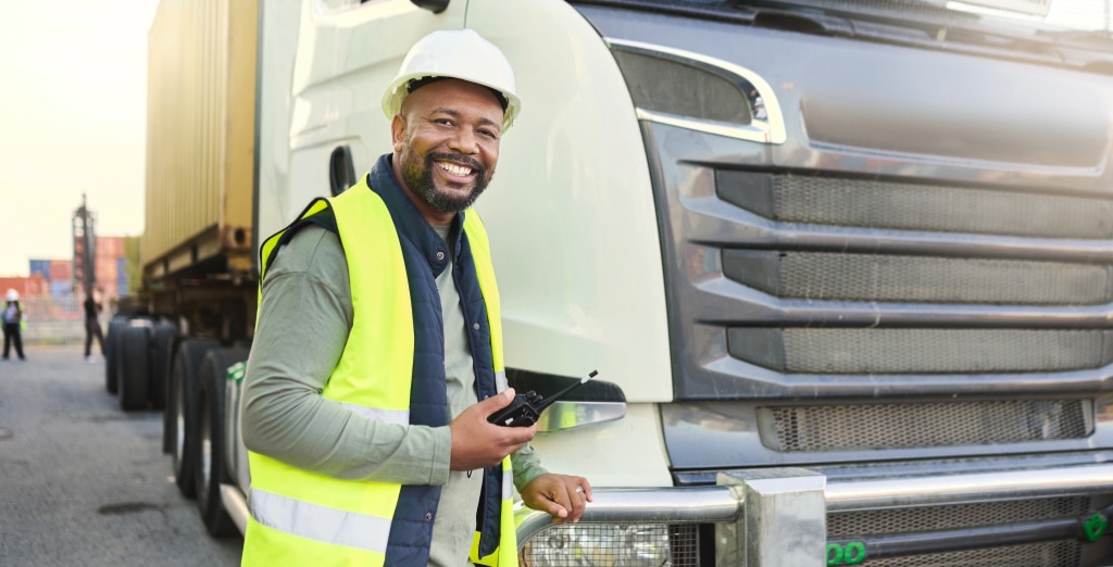 A smiling construction worker in a high-visibility vest and hard hat stands beside a parked truck, holding a walkie-talkie. The background, showcasing a large shipping container, reflects the importance of transport safety training in this bustling industrial setting.