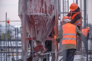 Construction workers in orange safety vests and helmets operate machinery on a building site, focusing on equipment maintenance. One worker handles a red concrete funnel, while others assist nearby amidst steel beams and rebar structures.