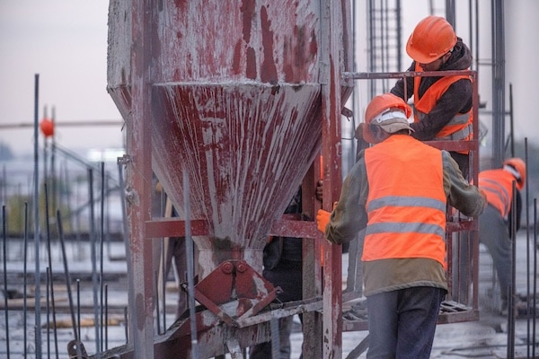 Construction workers in orange safety vests and helmets operate machinery on a building site, focusing on equipment maintenance. One worker handles a red concrete funnel, while others assist nearby amidst steel beams and rebar structures.