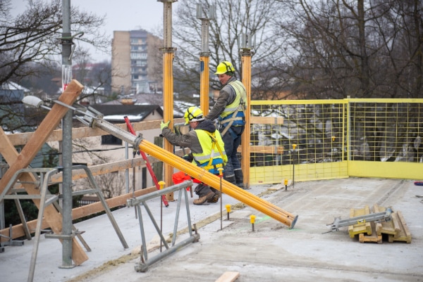 Two construction workers in safety gear are on a building site, diligently positioning wooden beams supported by metal scaffolding. With trees and a cityscape in the background, they focus on their task, mindful of preventing injuries under the brisk conditions. A yellow safety railing stands to the right.