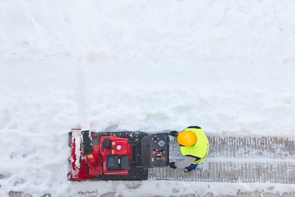 A person wearing a yellow safety vest and hard hat expertly operates a red snowblower, demonstrating snow and ice management by clearing a snowy path. Snow is piled on either side of the track being cleared, observed from above.