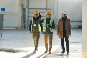 Three people in an industrial setting, two wearing yellow safety vests and hard hats, appear to be discussing best practices. The third person, in a brown coat and white hard hat, walks alongside them. They are inside a large, unfinished building.