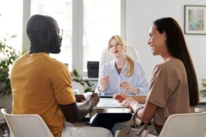 A diverse couple sits across a desk from a professional woman in an office setting, discussing workplace mental health. The woman at the desk speaks while the couple listens attentively. The room is bright with natural light, and plants are visible in the background, creating a calming environment.