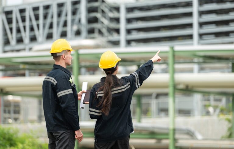 Two construction workers wearing yellow helmets and dark uniforms stand in front of an industrial facility. One holds rolled-up plans while the other points towards the building structure, appearing to discuss or inspect the site.