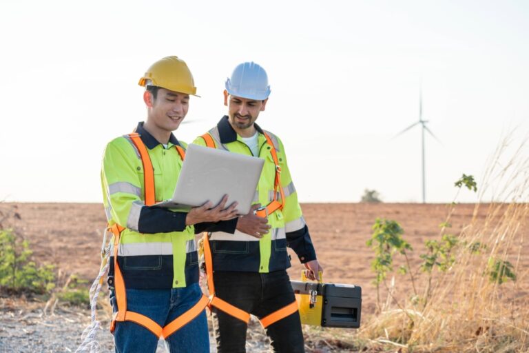 Two construction workers in safety gear and helmets stand outdoors. One holds a laptop, while the other carries equipment. A wind turbine is visible in the background. They appear to be in a rural area during daytime.