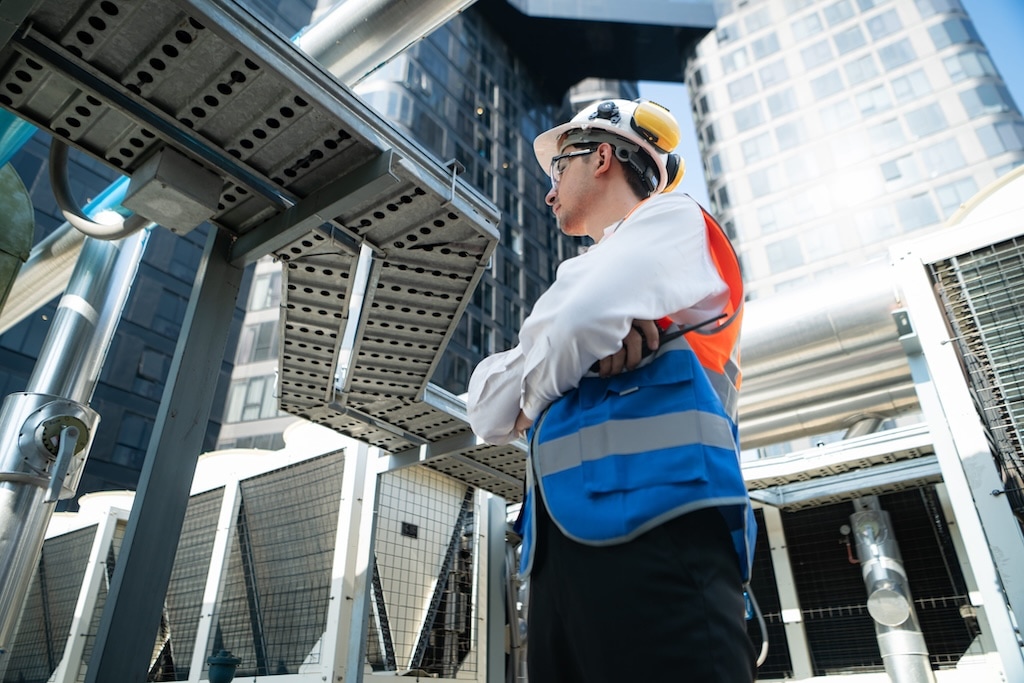 A construction worker in a white hard hat and reflective vest inspects industrial equipment on a rooftop. The worker stands with arms crossed, looking upward at a structure, with glass buildings in the background.