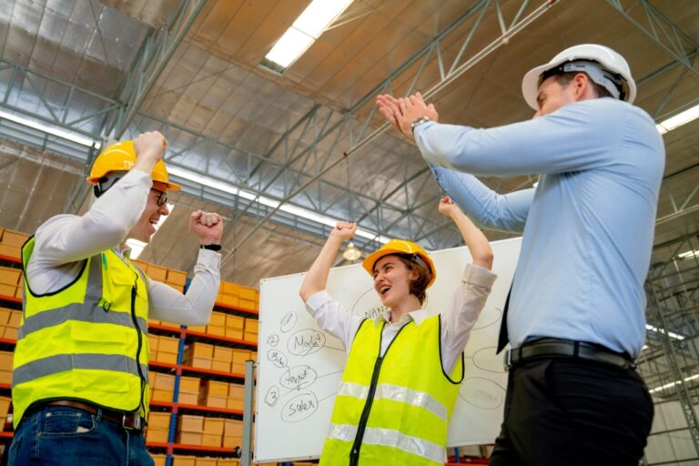 Three warehouse workers wearing safety vests and hard hats celebrate near a whiteboard with diagrams. They are in a spacious warehouse with stacks of boxes. The atmosphere is joyful and energetic.