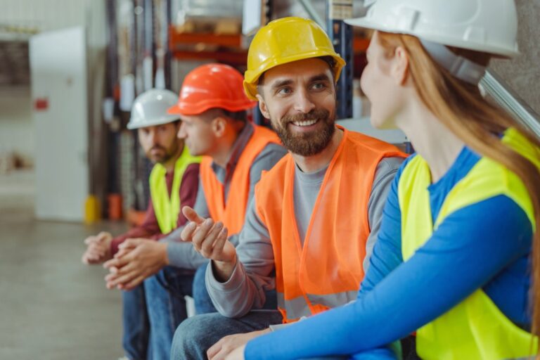 Four construction workers in safety vests and helmets sit on a step, engaged in conversation. The focus is on a bearded man in a yellow helmet talking to a woman in a white helmet. Other workers are seated in the background.