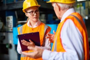 Two workers in safety vests and helmets have a discussion in an industrial setting. One holds a clipboard and appears attentive, while the other gestures with his hands. Machinery is visible in the background.