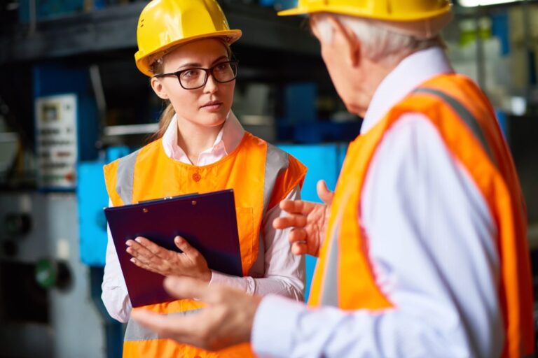 Two workers in safety vests and helmets have a discussion in an industrial setting. One holds a clipboard and appears attentive, while the other gestures with his hands. Machinery is visible in the background.