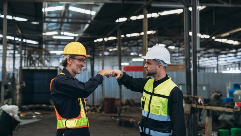 Two workers in hard hats and safety vests exchange a fist bump in a large industrial warehouse. They appear to be acknowledging each other amid various equipment and a high, open ceiling.