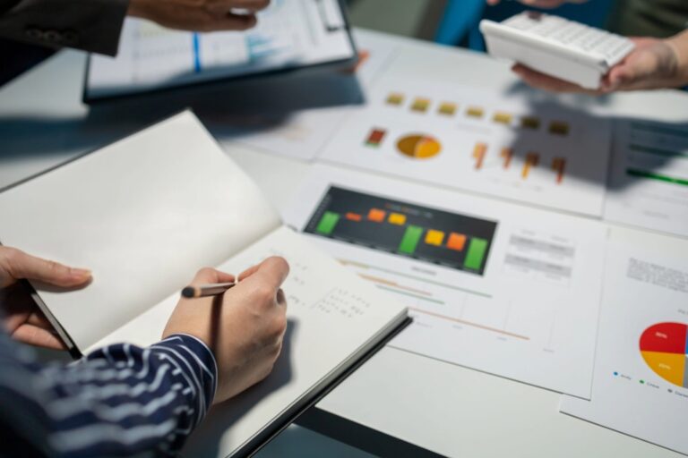 People collaborating on financial documents. One person writes in a notebook while another holds a calculator. Graphs and charts are spread across the table, indicating a focus on data analysis and business planning.