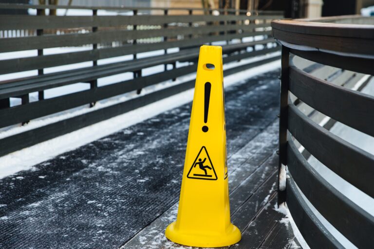 A yellow caution cone with an exclamation mark and slipping figure symbol stands on a snow-dusted walkway with dark wooden railings. The warning highlights slippery conditions during the winter months, alerting to potential slips, trips, and falls on the icy or snowy surface.