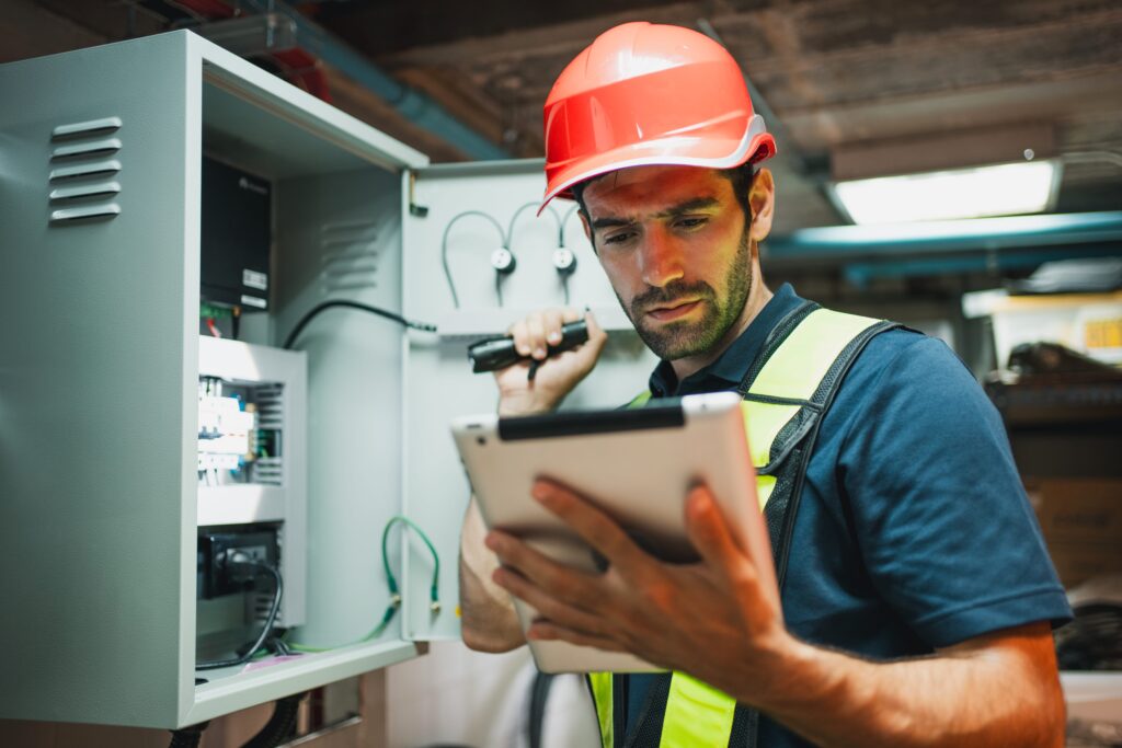 A construction worker in a red hard hat and yellow safety vest uses a flashlight to uncover hidden electrical hazards in an old building. With a tablet in hand, he focuses intently on the electrical panel, ensuring safety in this industrial environment.