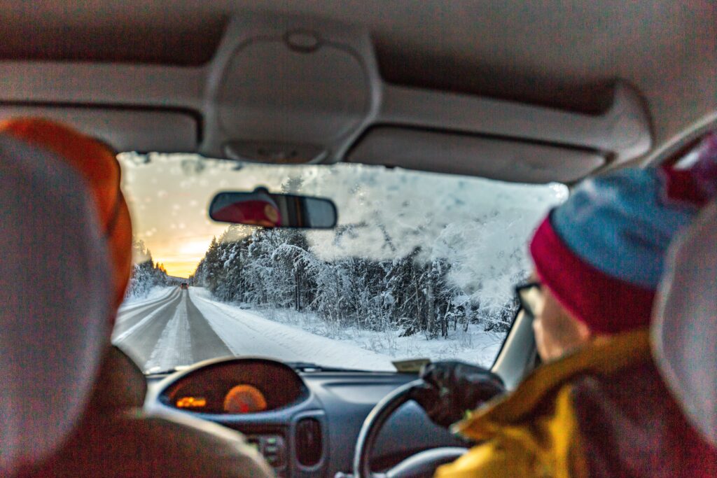 Two people in winter clothing navigate a snowy road flanked by frosty trees. The windshield, partially frosted, frames a sunrise or sunset. This serene, wintry scene underscores the importance of winter driving safety for all travelers on icy paths.