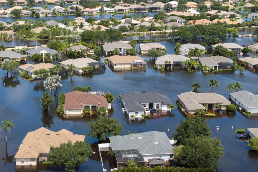 Aerial view of a suburban area heavily flooded, likely a result of recent winter storms. Numerous houses, surrounded by water, have only their roofs visible. Roads are submerged, and trees and shrubs are partially underwater. The scene reflects severe natural disasters.