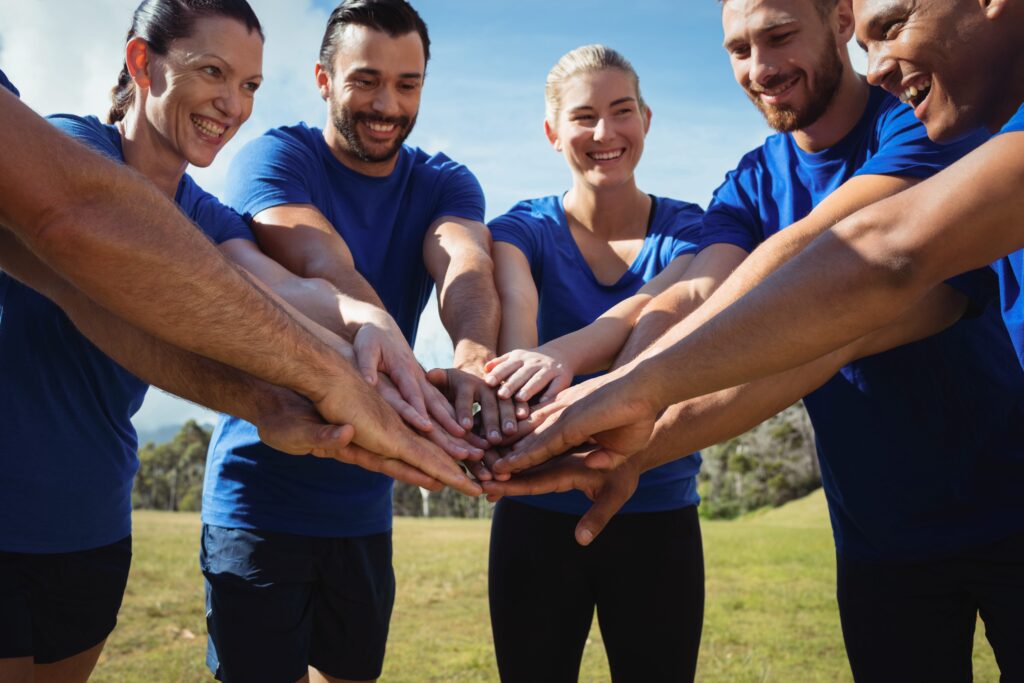 A group of people in blue shirts, standing outdoors with their hands joined in the center, smiling and looking at each other, embodies workplace wellness. They appear to be in a field with a clear sky in the background.