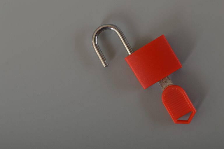 A small red padlock with a silver shackle is open, lying on a gray surface. The red body features a key in its base, illustrating the importance of Lockout Tagout Procedures to save lives.