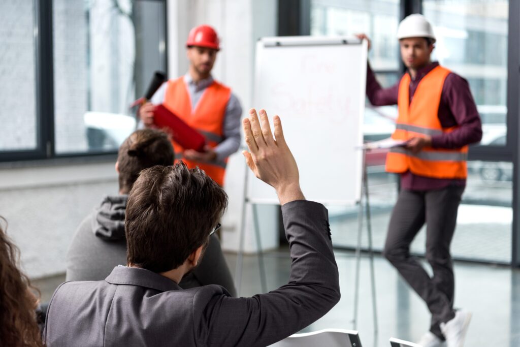 During a fire safety presentation, a man in the audience raises his hand as two presenters, clad in orange safety vests and hard hats, offer essential safety tips. One gestures towards the whiteboard while the other holds a clipboard, ensuring everyone learns how to maintain a safer workforce.