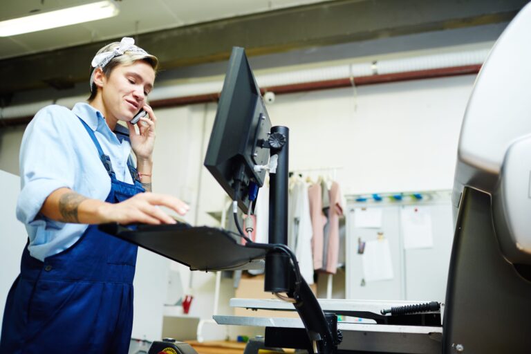 A person in blue overalls and a headscarf is multitasking at their workplace, using a computer and talking on the phone. They stand in an industrial setting with clothing and machinery, highlighting the importance of ergonomics in injury prevention.