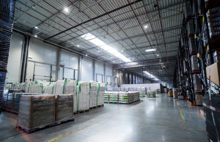 A spacious warehouse interior with high ceilings, filled with large bags and stacks of packaged goods on wooden pallets. Shelving units line the right side, and ergonomic lighting boosts productivity while illuminating the polished concrete floor.