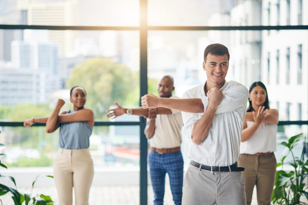 In a modern 2025 workplace, a diverse group of four people in business attire stretch their arms in an office with large windows. The man in front smiles at the camera, embracing techniques to reduce chronic work stress, while others focus on their stretches. Lush greenery is visible outside.