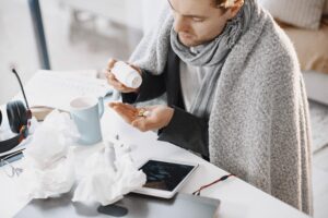 A person sits at a desk wrapped in a blanket, pouring pills into their hand. Tissue papers, a cup, headphones, and a tablet clutter the surface. They appear unwell, embodying the toll of seasonal illness as they focus on their medication while trying to protect themselves from further discomfort.
