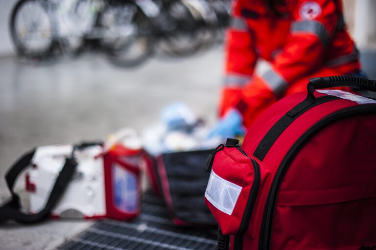 A person in a red uniform is kneeling and sorting through medical supplies from open emergency bags on the ground, embodying the spirit of workplace wellness programs. The focus is on a red backpack in the foreground, with blurred bicycles in the background.