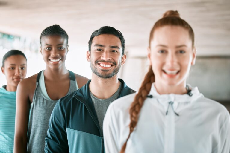 A diverse group of four people stands in a line outdoors, smiling at the camera, perhaps in celebration of their workplace wellness program. They are wearing athletic clothing, suggesting they are about to engage in physical activity. The scene is set in an urban environment.