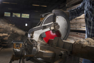 A person in a plaid shirt operates a circular saw, maintaining focus on safety to prevent hazards. Sawdust flies as the red blade spins, but care is taken to manage combustible dust. Stacks of wooden planks in the background suggest an active workshop setting.