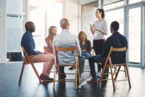 A group of six people sits in a circle on wooden chairs in a bright room. One stands, gesturing as they speak, while the others listen attentively. The scene suggests a meeting focused on fostering a mentally healthy workplace through effective safety programs.
