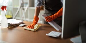 A person in orange gloves ensures cleanliness as they meticulously clean a desk. Holding a white keyboard in one hand and using a cloth with the other, they show keen accountability. A spray bottle and computer monitor stand ready, underscoring the importance of housekeeping safety.