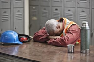 A construction worker in an orange safety vest rests their head on crossed arms atop a table, managing fatigue. Nearby, a blue hard hat, ear protection, thermos, and cup sit untouched. Lockers loom in the background as a reminder of the importance of vigilance to avoid workplace accidents.