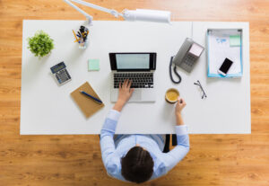 A person sits at a desk using a laptop, surrounded by a notebook, calculator, and cup of coffee. With an eye on workplace safety, the clean workspace includes a plant, lamp, phone, and glasses neatly arranged on the wooden floor visible beneath.