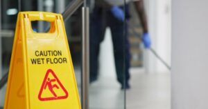 Yellow caution sign with "Caution Wet Floor" text and slipping figure icon. In the background, a person wearing blue gloves is cleaning a floor with a mop near glass doors. The focus is on the sign.
