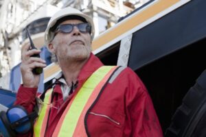 A person in a red jacket and yellow safety vest, wearing a white hard hat and sunglasses, holds a walkie-talkie. They are looking up, practicing sun safety to protect skin and eyes. The background shows partial industrial equipment.