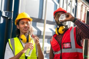 Two construction workers wearing safety vests, helmets, and ear protection stand by a shipping container in the extreme heat. Embracing the buddy system for safety, one drinks from a water bottle while the other holds theirs, both enjoying a well-earned break.